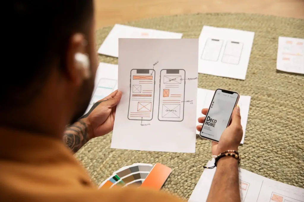 Imagem ilustrativa sobre a arquitetura de site. Mostra uma pessoa segurando um protótipo de um site em papel e um smartphone que exibe uma aplicação. O fundo contém esboços e elementos de design.